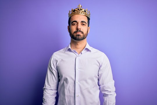 Young handsome man with beard wearing golden crown of king over purple background Relaxed with serious expression on face. Simple and natural looking at the camera.
