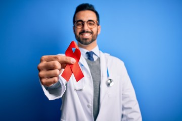 Young handsome doctor man with beard wearing stethoscope holding red hiv ribbon with a happy face...