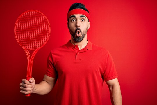 Young handsome sportsman with beard playing tennis using racket over red background scared in shock with a surprise face, afraid and excited with fear expression