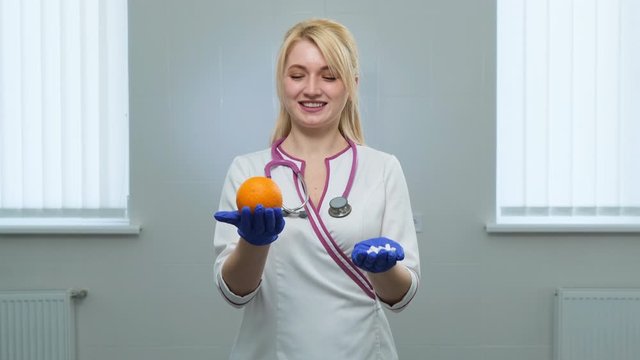 Caucasian young doctor in white medical coat, violet stethoscope holding a white pills with left hand and orange in rigth hand in blue gloves. Choice of medicine, pill or fruits and vitamins concept