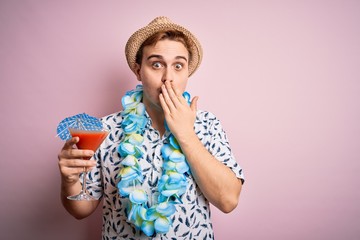 Young redhead tourist man on vacation wearing hat and hawaiian lei drinking cocktail covering mouth...