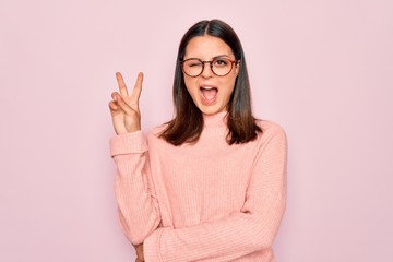 Young beautiful brunette woman wearing casual sweater and glasses over pink background smiling with happy face winking at the camera doing victory sign. Number two.