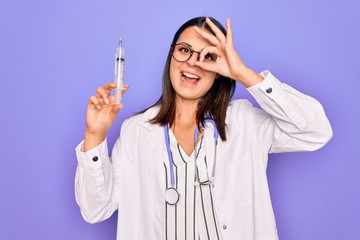 Young beautiful brunette doctor woman wearing stethoscope holding syringe vaccination smiling happy doing ok sign with hand on eye looking through fingers