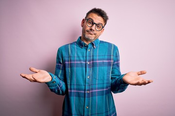 Young handsome man wearing casual shirt and glasses standing over isolated pink background clueless...