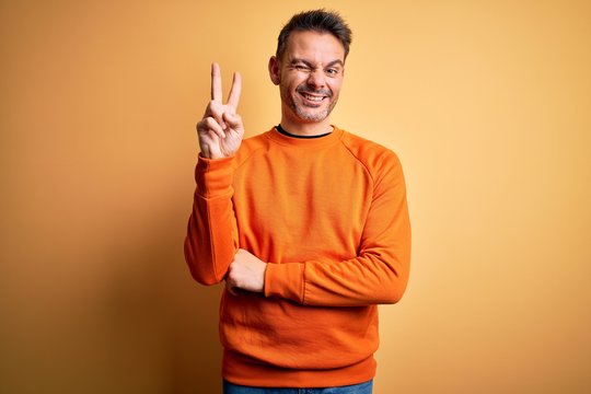 Young handsome man wearing orange casual sweater standing over isolated yellow background smiling with happy face winking at the camera doing victory sign. Number two.