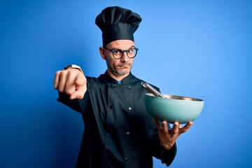 Young handsome chef man wearing cooker uniform and hat mixing using whisk and bowl pointing with finger to the camera and to you, hand sign, positive and confident gesture from the front