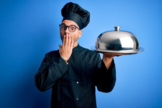 Young handsome chef man wearing uniform and hat holding waiter tray with dome cover mouth with hand shocked with shame for mistake, expression of fear, scared in silence, secret concept