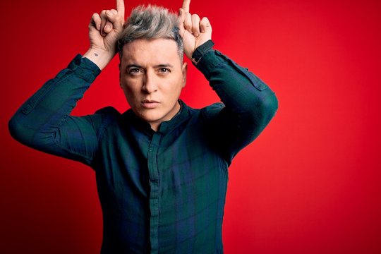 Young handsome modern man wearing elegant green shirt over red isolated background doing funny gesture with finger over head as bull horns