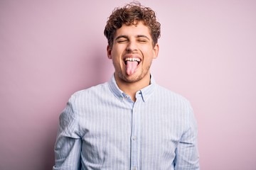 Young blond handsome man with curly hair wearing striped shirt over white background sticking tongue out happy with funny expression. Emotion concept.