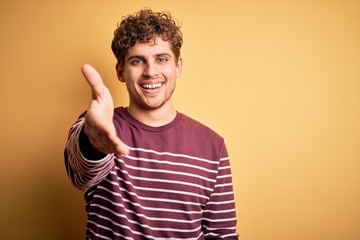 Young blond handsome man with curly hair wearing casual striped sweater smiling friendly offering handshake as greeting and welcoming. Successful business.
