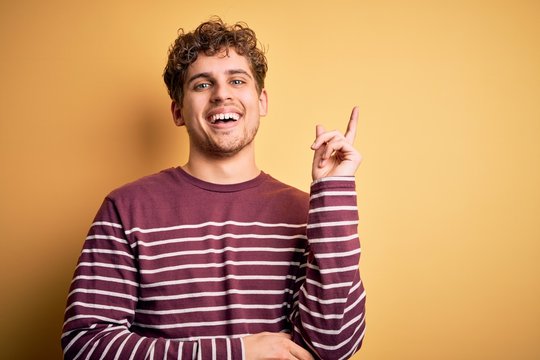Young blond handsome man with curly hair wearing casual striped sweater with a big smile on face, pointing with hand and finger to the side looking at the camera.