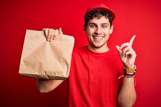 Young blond delivery man with curly hair wearing cap holding paper bag with food surprised with an idea or question pointing finger with happy face, number one