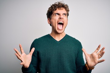 Young blond handsome man with curly hair wearing green sweater over white background crazy and mad shouting and yelling with aggressive expression and arms raised. Frustration concept.