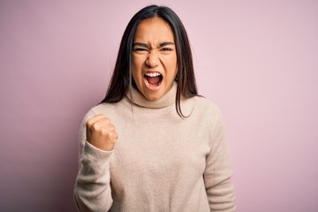 Young beautiful asian woman wearing casual turtleneck sweater over pink background angry and mad raising fist frustrated and furious while shouting with anger. Rage and aggressive concept.