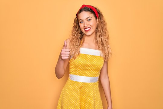 Beautiful blonde pin-up woman with blue eyes wearing diadem standing over yellow background doing happy thumbs up gesture with hand. Approving expression looking at the camera showing success.