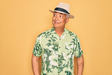 Middle age senior grey-haired man wearing summer hat and floral shirt on beach vacation smiling looking to the side and staring away thinking.