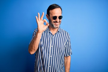 Naklejka premium Middle age hoary man wearing striped shirt and sunglasses over isolated blue background smiling positive doing ok sign with hand and fingers. Successful expression.