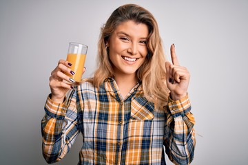 Young beautiful blonde woman drinking glass of healthy orange juice over white background surprised with an idea or question pointing finger with happy face, number one