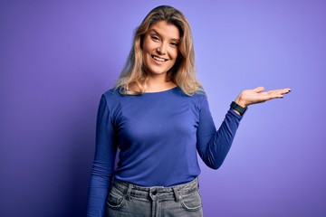 Young beautiful blonde woman wearing casual t-shirt over isolated purple background smiling cheerful presenting and pointing with palm of hand looking at the camera.