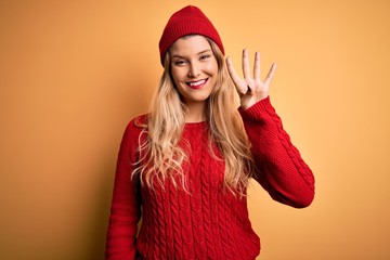 Young beautiful blonde woman wearing casual sweater and wool cap over white background showing and pointing up with fingers number four while smiling confident and happy.