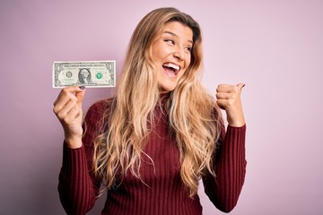 Young beautiful blonde woman holding one dollar banknote over isolated pink background pointing and showing with thumb up to the side with happy face smiling
