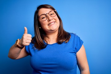 Beautiful brunette plus size woman wearing casual t-shirt over isolated blue background doing happy thumbs up gesture with hand. Approving expression looking at the camera showing success.