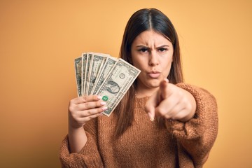 Young beautiful woman holding dollars standing over isolated orange background pointing with finger to the camera and to you, hand sign, positive and confident gesture from the front