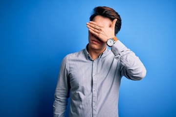 Young handsome chinese man wearing casual shirt standing over isolated blue background covering eyes with hand, looking serious and sad. Sightless, hiding and rejection concept