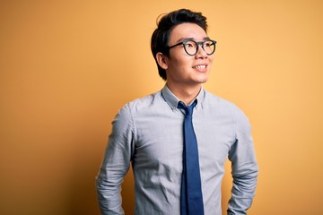 Young handsome chinese businessman wearing glasses and tie over yellow background looking away to...