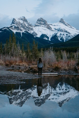 Fototapeta na wymiar Tourist woman looking at iconic the Three Sisters mountain peaks reflecting in in Policeman's Creek calm water while sunset. Canmore, Alberta, Canada.