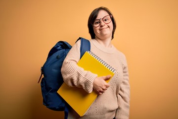 Young down syndrome student woman wearing glasses and school back holding books from library with a happy face standing and smiling with a confident smile showing teeth