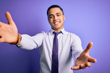 Young brazilian businessman wearing elegant tie standing over isolated purple background looking at the camera smiling with open arms for hug. Cheerful expression embracing happiness.