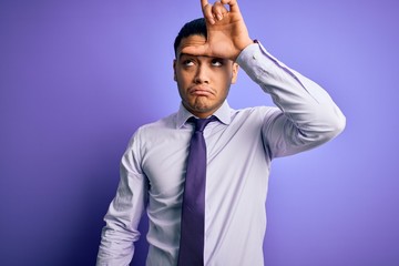 Young brazilian businessman wearing elegant tie standing over isolated purple background making fun of people with fingers on forehead doing loser gesture mocking and insulting.
