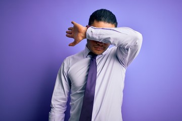 Young brazilian businessman wearing elegant tie standing over isolated purple background covering eyes with arm, looking serious and sad. Sightless, hiding and rejection concept