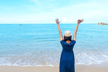 Fototapeta na wymiar Asian young teenager girl wearing sun hat & cute navy blue dress holding mobile phone or smartphone enjoying & admiring of deep blue sea view with soft calm waves touching tropical summer sand beach