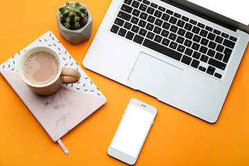 Modern mobile phone with laptop, cup of coffee and notebook on color background