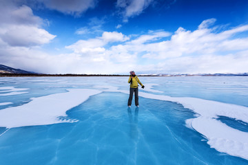 A girl in a yellow jacket and ice skates stands on the blue lake of lake Baikal and looks at the sky