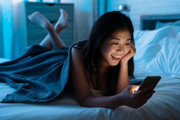 Happy asian japanese teen lying in front in bed at home with smartphone. Young joyful woman watching funny video on mobile phone in bedroom at night and laughing. relax girl shopping online indoors