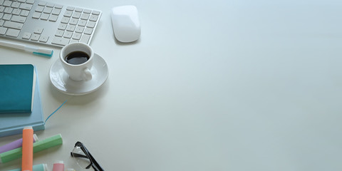 Top view image of coffee cup putting on white working desk that surrounded by wireless mouse, keyboard, marker pens, eye glasses, notebook, note and pen.