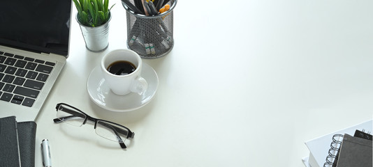 Photo of ceramic coffee cup with small dish putting on white working desk that surrounded by black empty screen computer laptop, potted plant, pen, pencil holder, document file, notebook and glasses.