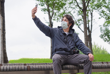 Fototapeta premium Young tourist with surgical mask in video call with his smartphone, photographer with health care mask in urban exploration take a selfie, sitting on a bench in a city in outbreak of Covid19