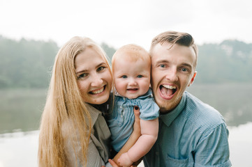 Fototapeta premium Happy young family near lake, pond. Family enjoying life together at meadow. People having fun in nature. Family bonds outside. Mother, father, child of three smiling while spending free time outdoors