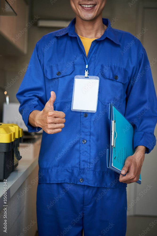 Wall mural Smiling young repairman in blue uniform holding clipboard and showing thumbs-up