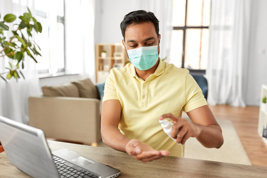 quarantine, remote job and pandemic concept - happy indian man wearing face protective medical mask for protection from virus disease with laptop computer using hand sanitizer at home office