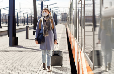 Fototapeta na wymiar health, safety and pandemic concept - young woman in protective face mask with travel bag walking along empty railway station