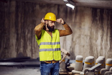 Young bearded worker in vest putting his helmet on head while standing inside building in construction process.