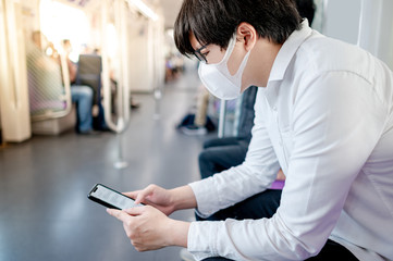 Obraz na płótnie Canvas Asian man wearing surgical face mask using smartphone on skytrain or urban train. Coronavirus (COVID-19) outbreak prevention in public transportation. Social distancing for pandemic protection