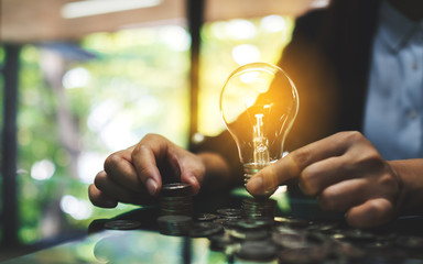 Businesswoman holding lightbulb while stacking coins on table for saving energy and money concept