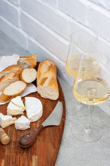 Two glasses of white wine, camemebert, cheese knife and baguette on wooden board.