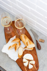 Two glasses of white wine, camemebert, cheese knife and baguette on wooden board.